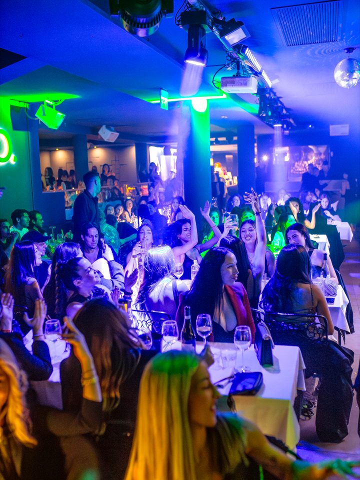 La sala piena durante la serata: ospiti ai tavoli con le braccia alzate sotto le luci blu